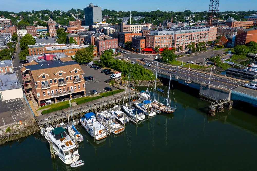 Aerial view of South Norwalk waterfront with boats docked in a marina, nearby buildings, and a bridge over the water.