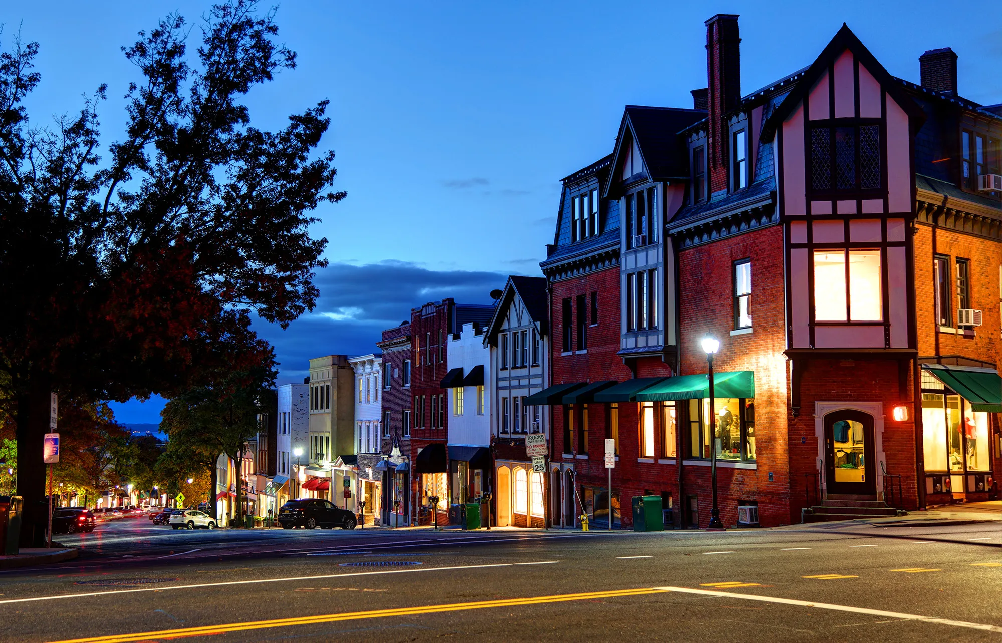 Greenwich real estate vibrant downtown street at dusk with historic Tudor brick buildings, glowing storefronts, and lamp posts