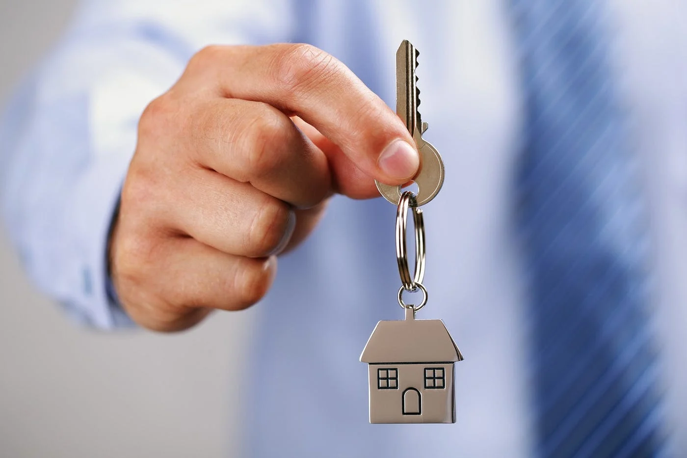 Hand holding a house key with a small house-shaped keychain in front of a blurred background.