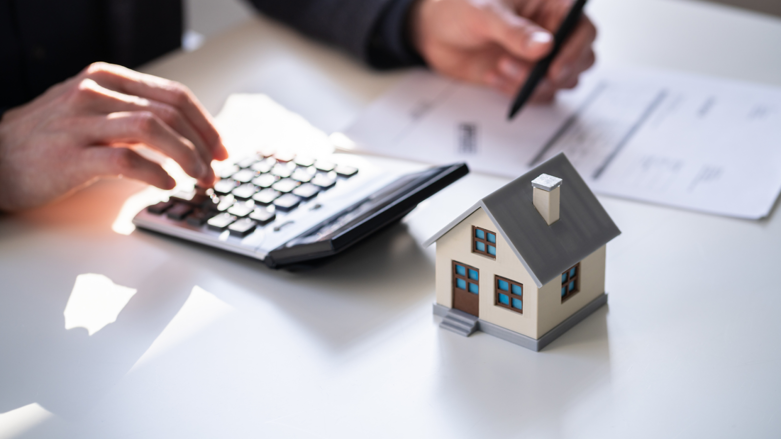 Hands working on paperwork with a calculator next to a small house model on a desk.