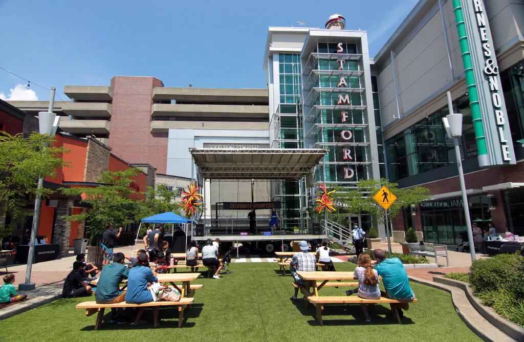 Outdoor plaza in Stamford with people seated at picnic tables watching a small stage performance in a mixed-use downtown area.