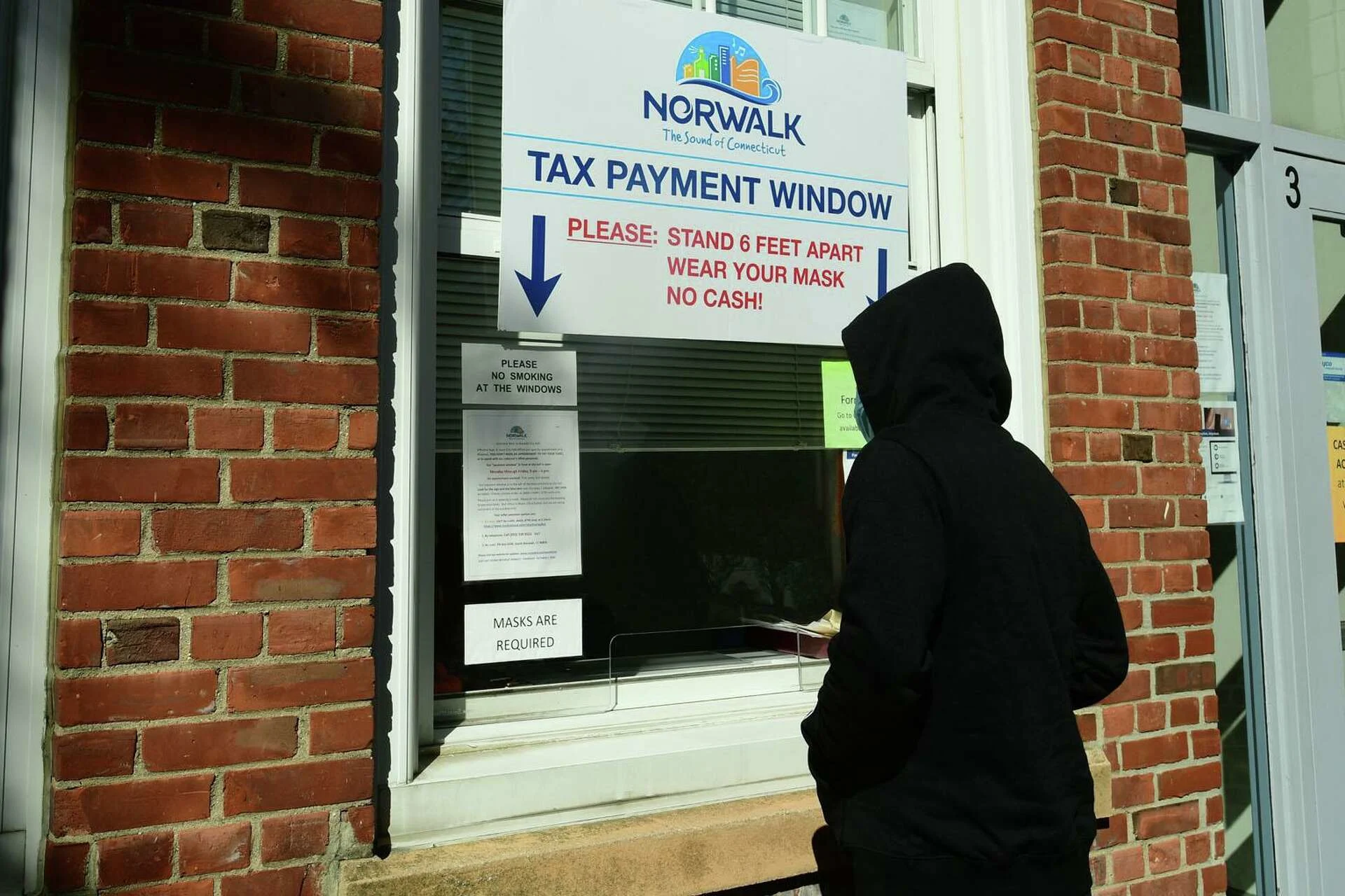 Person in a hoodie standing at a Norwalk “Tax Payment Window” counter outside a brick building