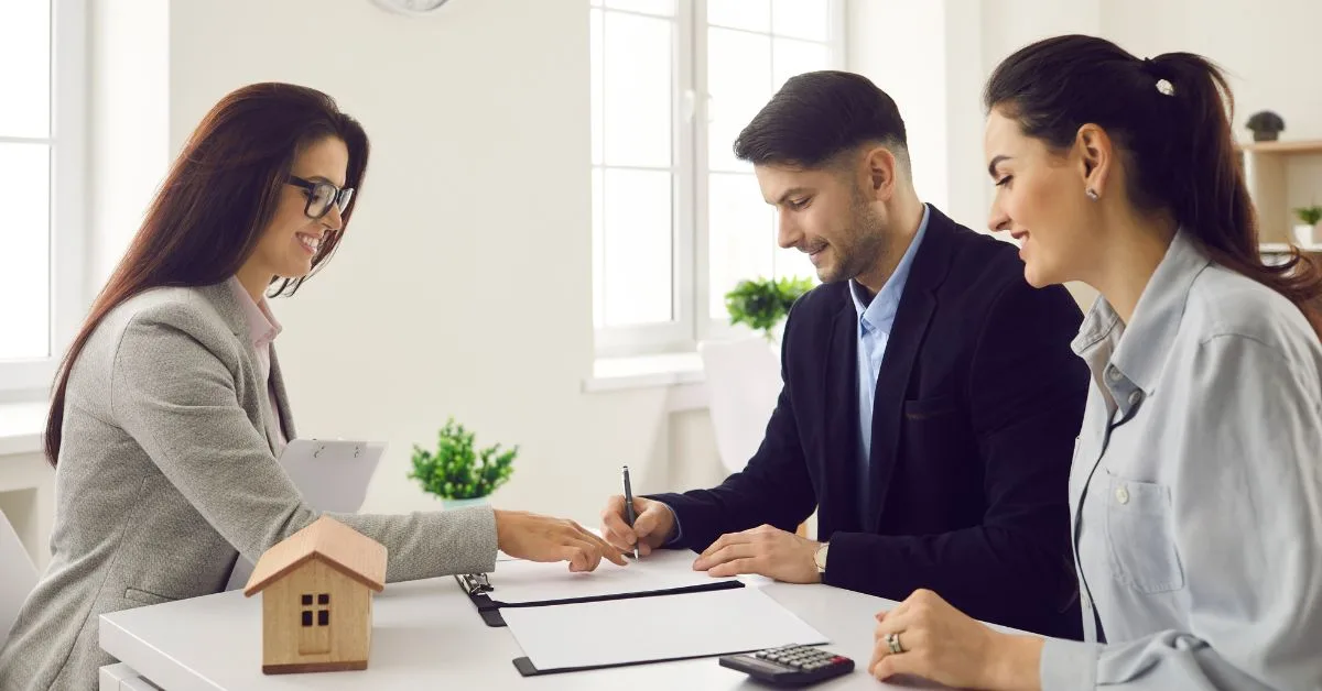 Real estate agent meeting with a couple signing documents beside a small house model.