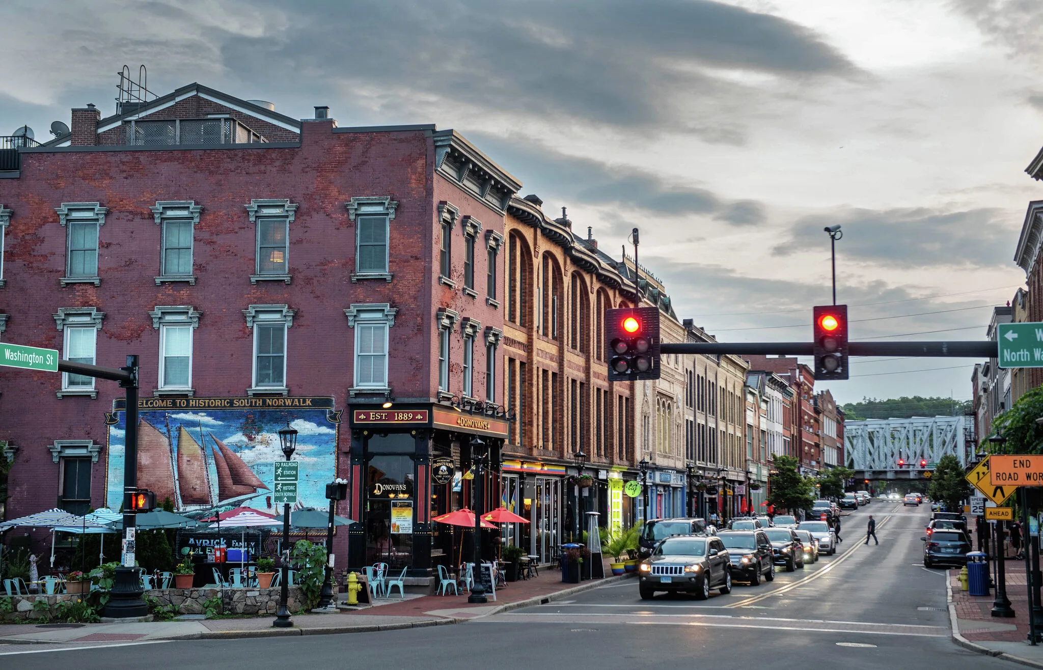 Street view of South Norwalk with brick buildings, restaurants, traffic, and cars waiting at a red light near an intersection.